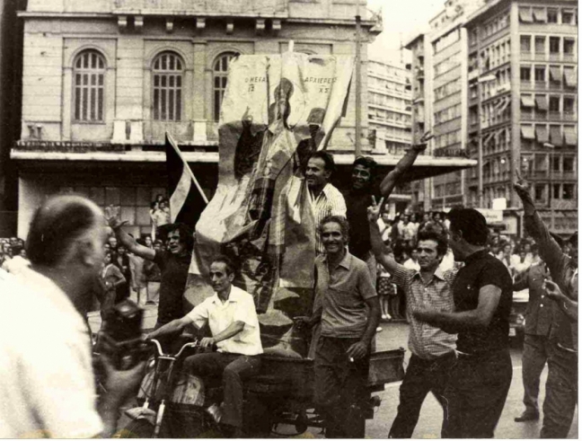 24 Ioυλίου 1974, Αθήνα, Ομόνοια. Πολίτες με σημαίες και μια τεράστια εικόνα του «Μεγάλου Αρχιερέως» πανηγυρίζουν την επιστροφή της δημοκρατίας και του Κωνσταντίνου Καραμανλή.