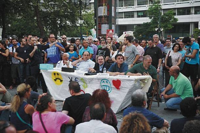 Τραπεζάκι συνειδητοποίησης των «Αγανακτισμένων» και κατανόησης των «βλαβερών συνεπειών του μνημονίου», το καλοκαίρι του 2011, στην πλατεία Συντάγματος. Από τους πέντε ομιλητές (από αριστερά: ένας άγνωστος, Καζάκης, Κατρούγκαλος, Τσακαλώτος, Βαρουφάκης), οι τρεις τελευταίοι είναι υπουργοί της κυβέρνησης Τσίπρα.