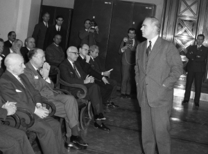 1962, Αθήνα. Ο πρωθυπουργός Κωνσταντίνος Καραμανλής επισκέπτεται την Τράπεζα της Ελλάδος, προκειμένου να ενημερωθεί για τις οικονομικές εξελίξεις της χώρας. Σε αίθουσα όπου θα προβληθούν τα οικονομικά διαγράμματα, τον πρωθυπουργό περιμένουν διάφοροι οικονομικοί παράγοντες, μεταξύ των οποίων ο Ξενοφών Ζολώτας (τρίτος από αριστερά).  